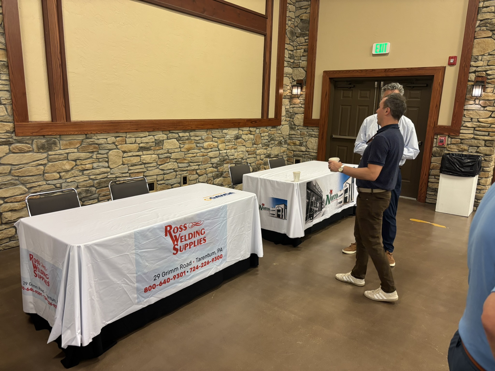 Two men converse beside vendor tables covered with Ross Welding Supplies banners in a stone-walled event room.