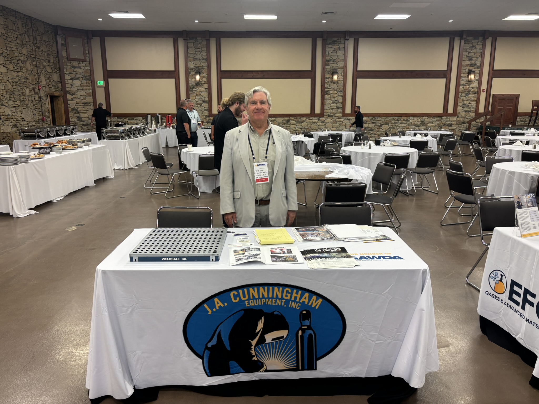 Man in a light gray blazer and badge stands at a banquet table in a large event hall with buffet stations along the left wall.
