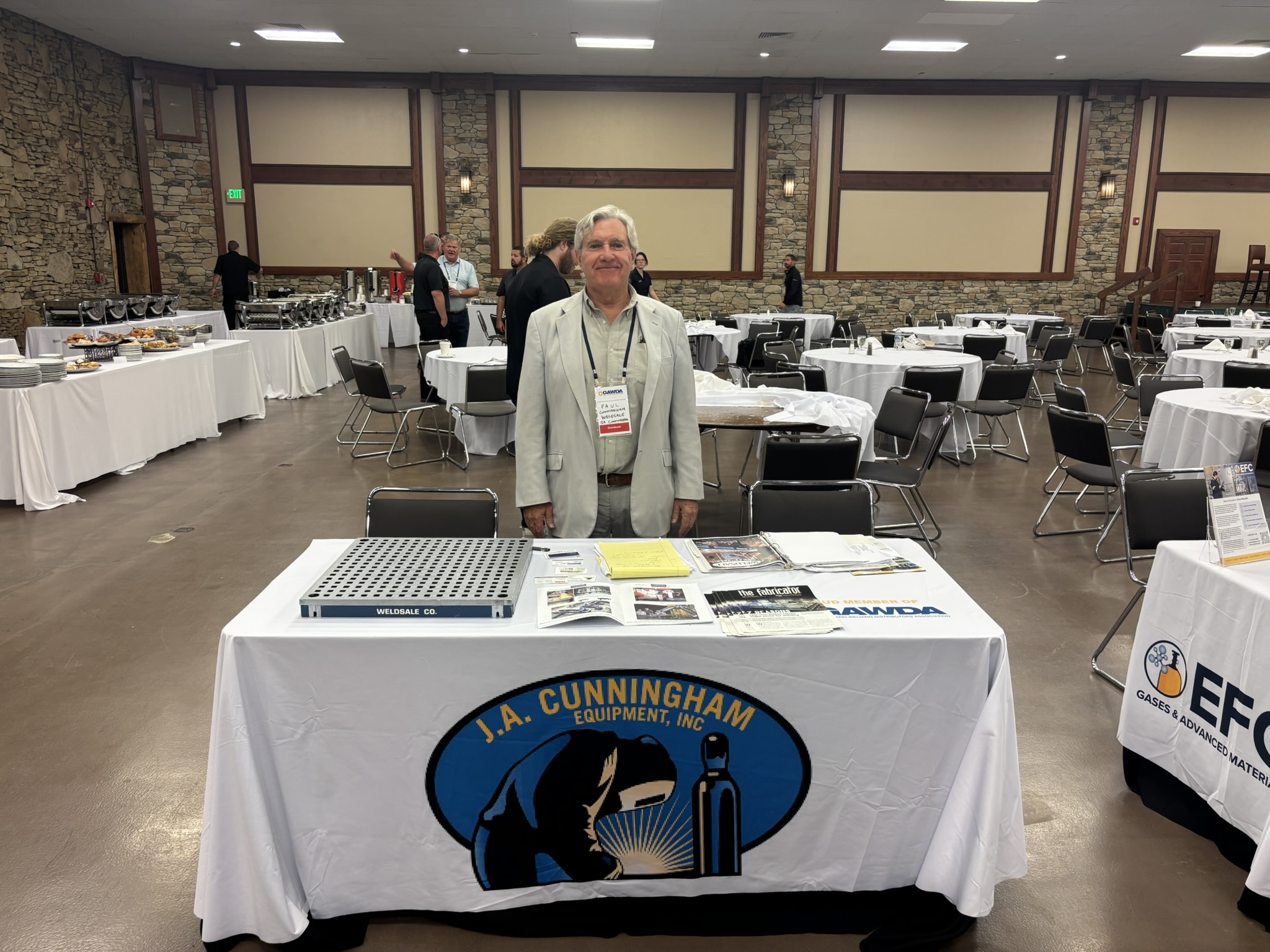 Man in a light gray suit with a name badge standing at a convention table in a banquet hall with rounds of tables and food stations along the back wall.