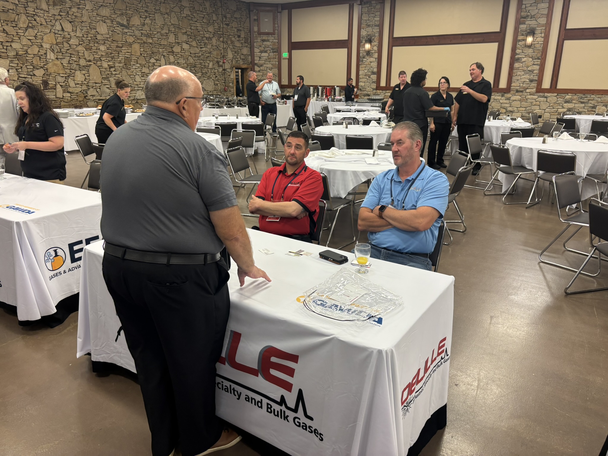 Banquet hall with a stone wall, round tables covered in white linens, and people standing and talking; foreground shows a man in a gray shirt speaking to two seated men in red and blue polos.