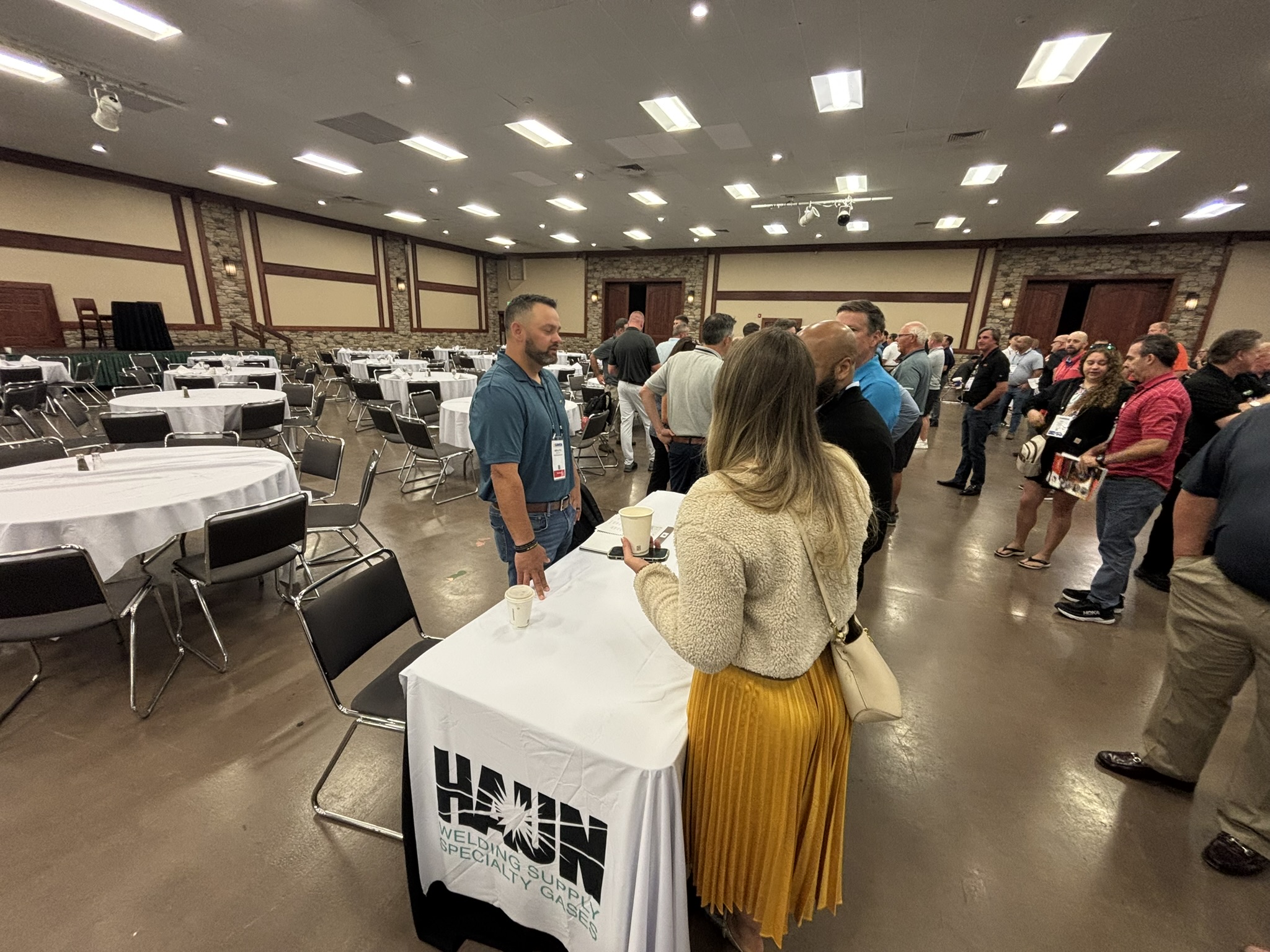 Line of attendees at a trade show or conference, standing in a queue near a vendor booth with a white-covered table and banner in a banquet-style hall.