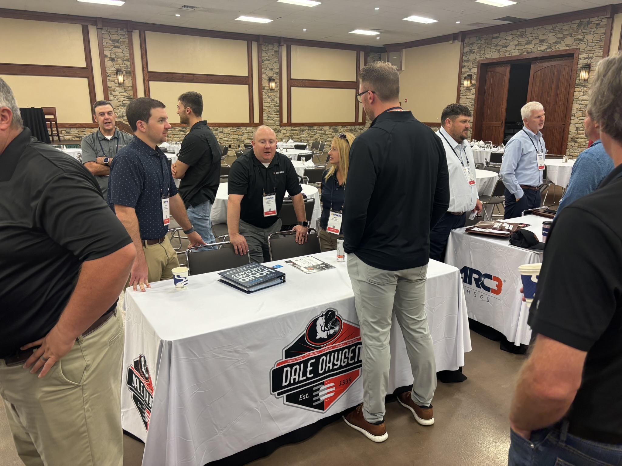 Group of professionals wearing name badges talk and mingle around tables in a conference room with stone walls and wood paneling; Dale Oxygen banners are visible on table covers and displays.