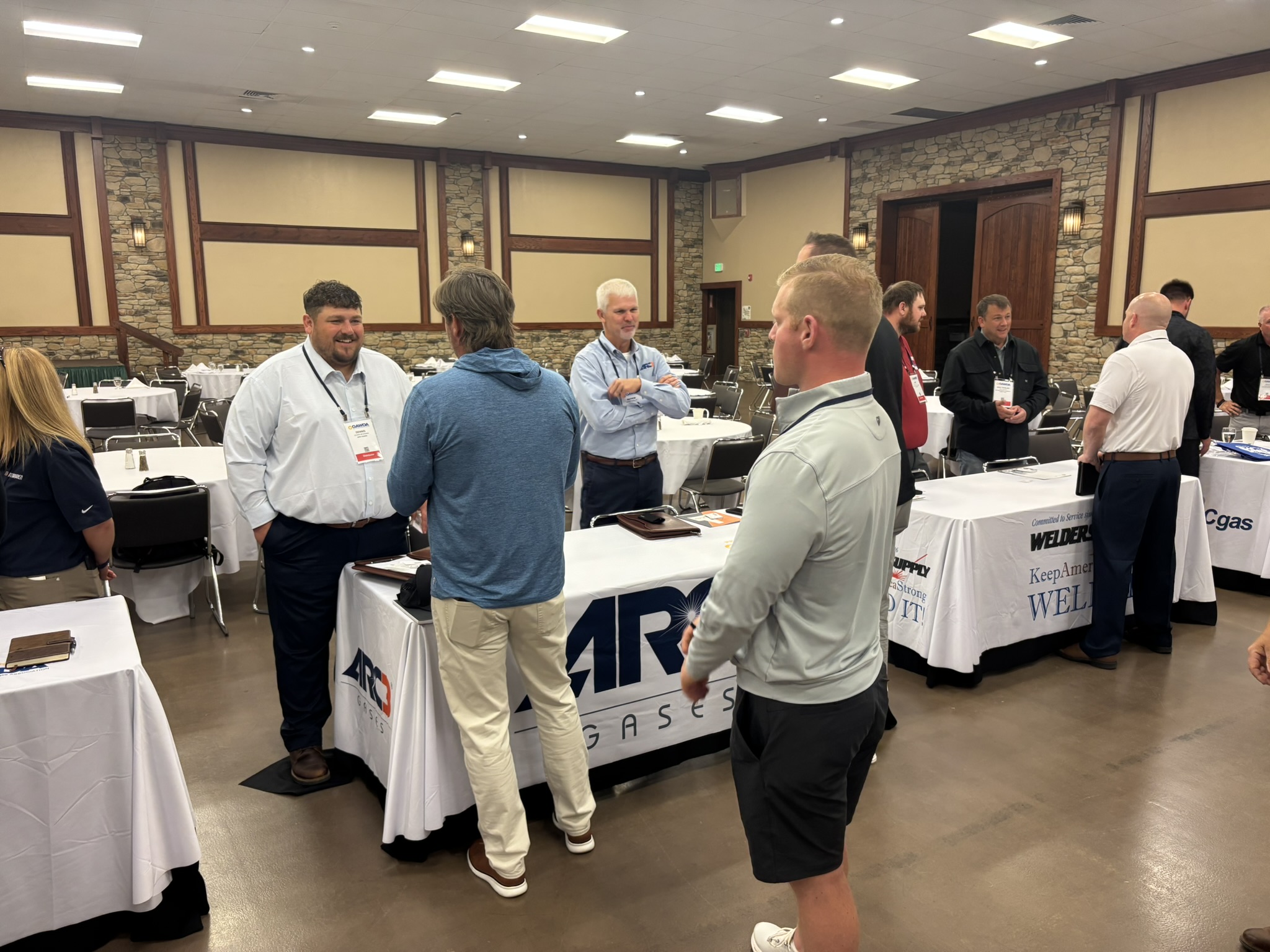 Group of men in business casual talking and networking around a vendor booth in a banquet-style conference room with round tables and white cloths.
