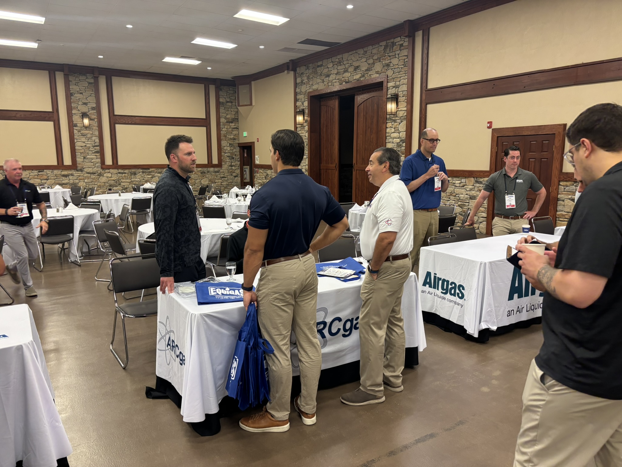 Men in business casual talk in a banquet hall with round tables, white tablecloths, and Airgas banners visible on the table covers.