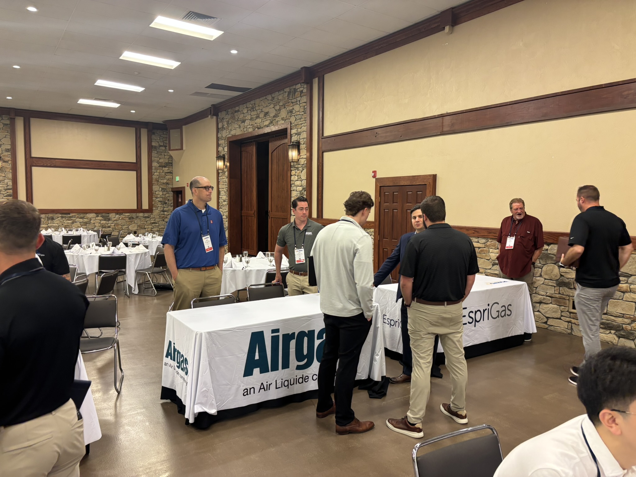 Group of attendees in a banquet-style room around a booth with Airgas banners, engaged in conversation during a corporate event.