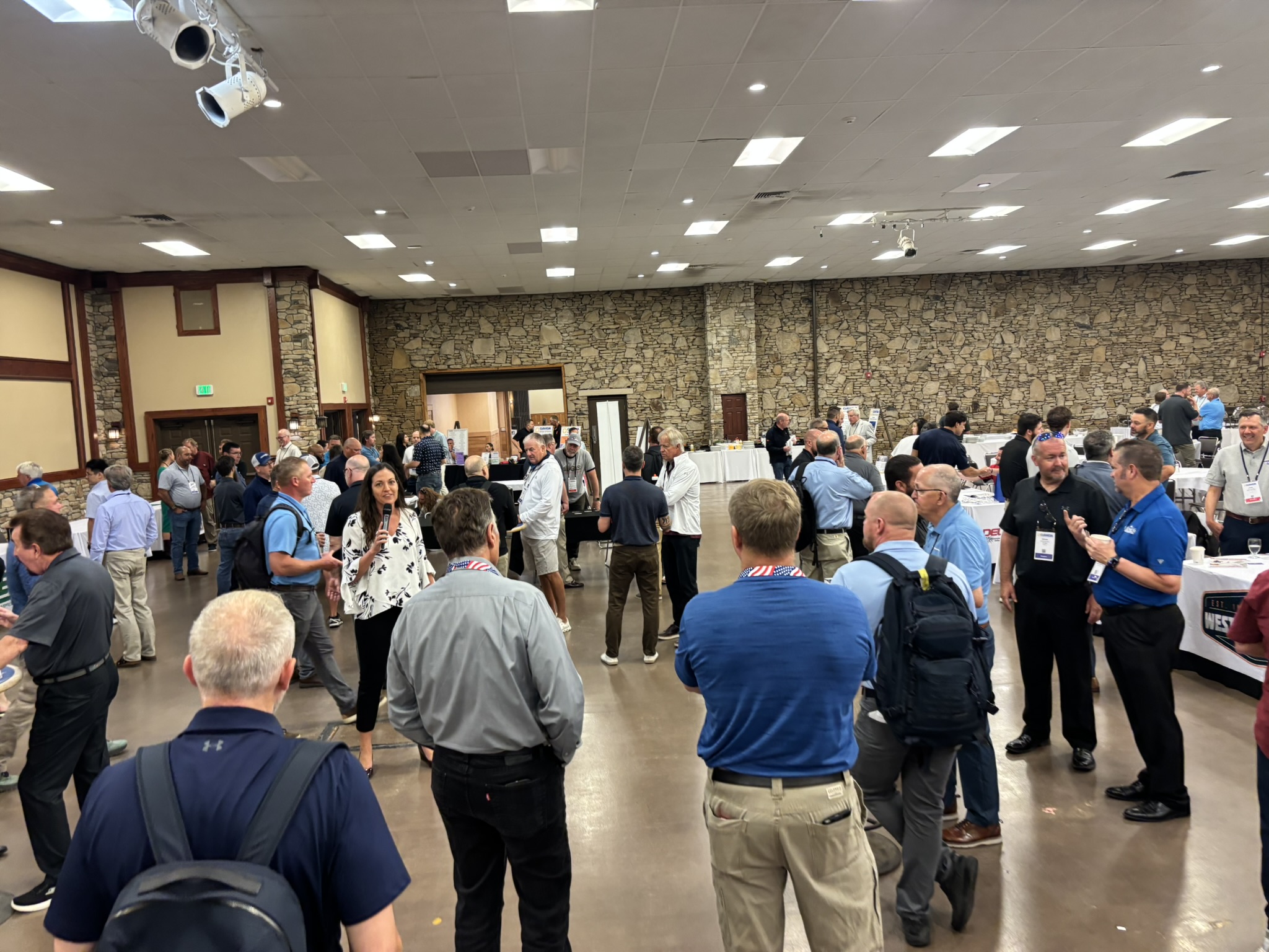 Large conference hall crowded with people standing and talking around round tables at a networking event.