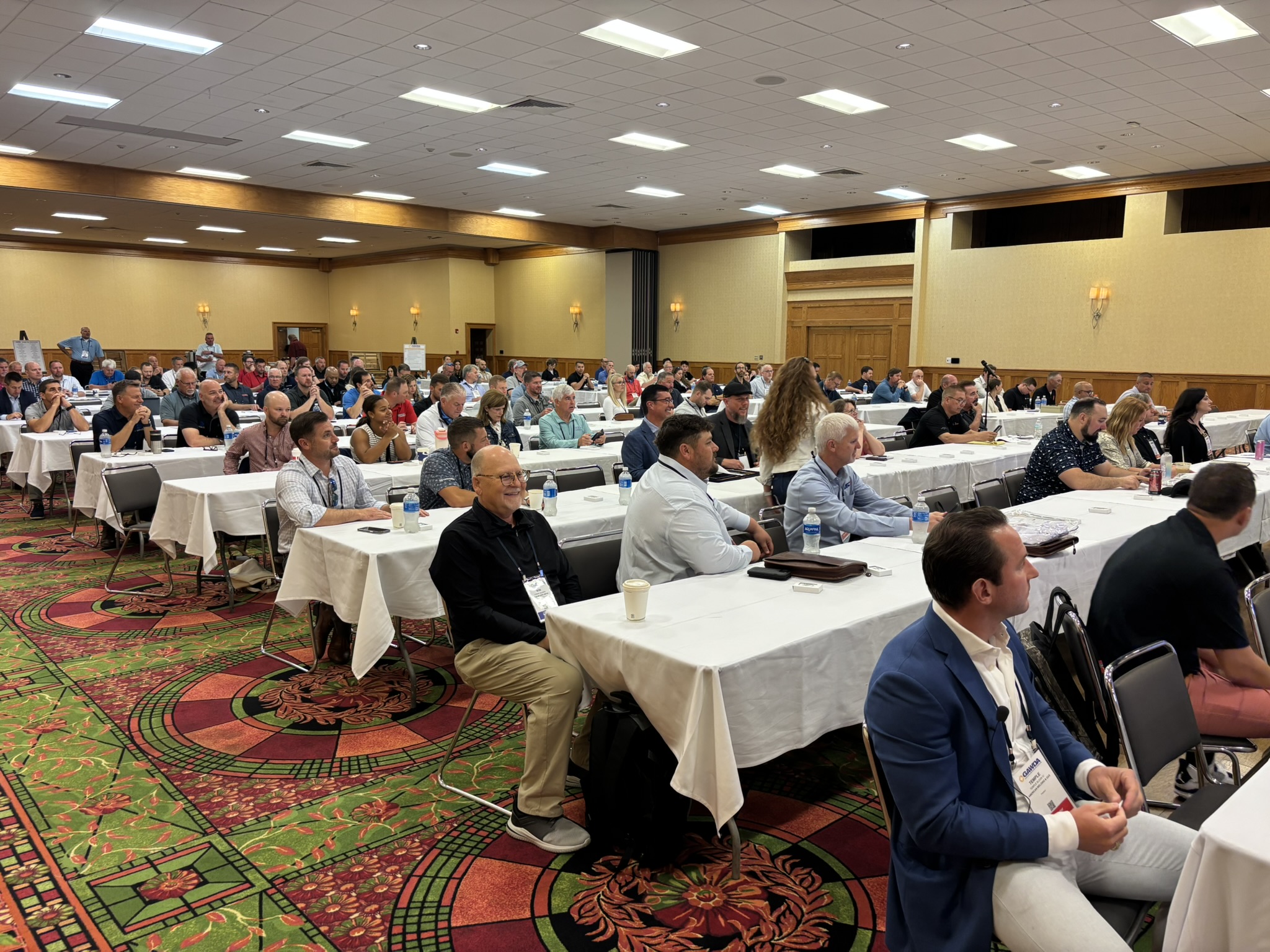 Conference room full of attendees seated at long white-table desks, listening to a presentation.