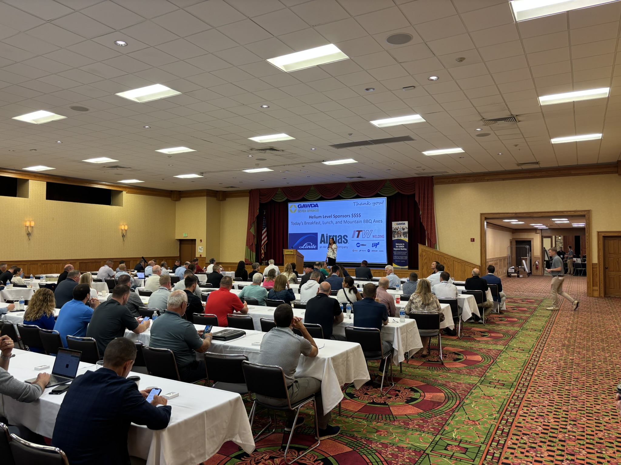 Conference hall filled with attendees seated at long tables facing a stage with a blue screen banner and speakers.