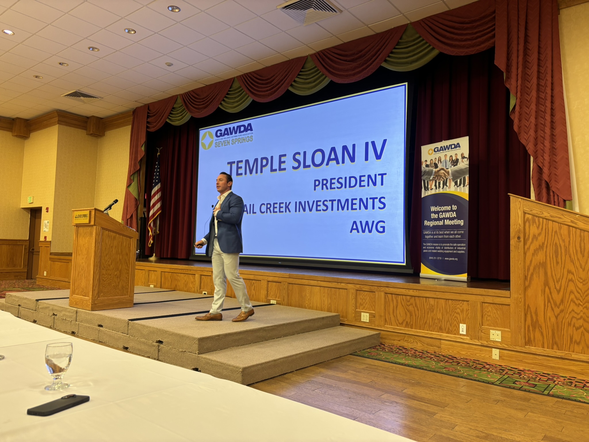 Male presenter in a blue blazer speaks on a stage with a large screen showing 'TEMPLE SLOAN IV' and 'PRESIDENT' text; stage has a podium, American flag, and red curtains in a conference room.