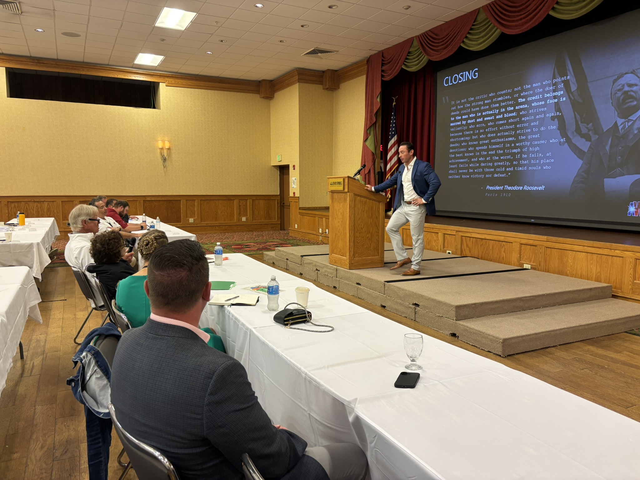Speaker in a blue blazer delivers closing remarks at a podium on a stage with a large projection screen nearby showing the word 'CLOSING' and a portrait, in a conference room with attendees at long tables.