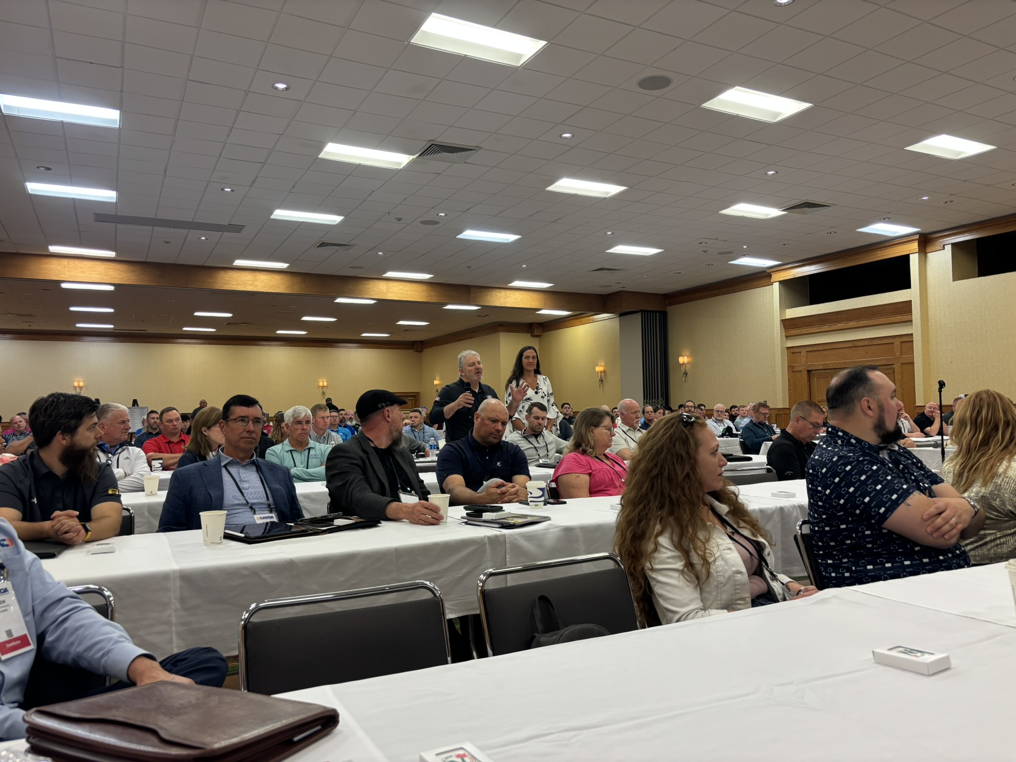 Large conference room filled with people seated at long tables listening to a speaker in a meeting setting, ceiling lights overhead.