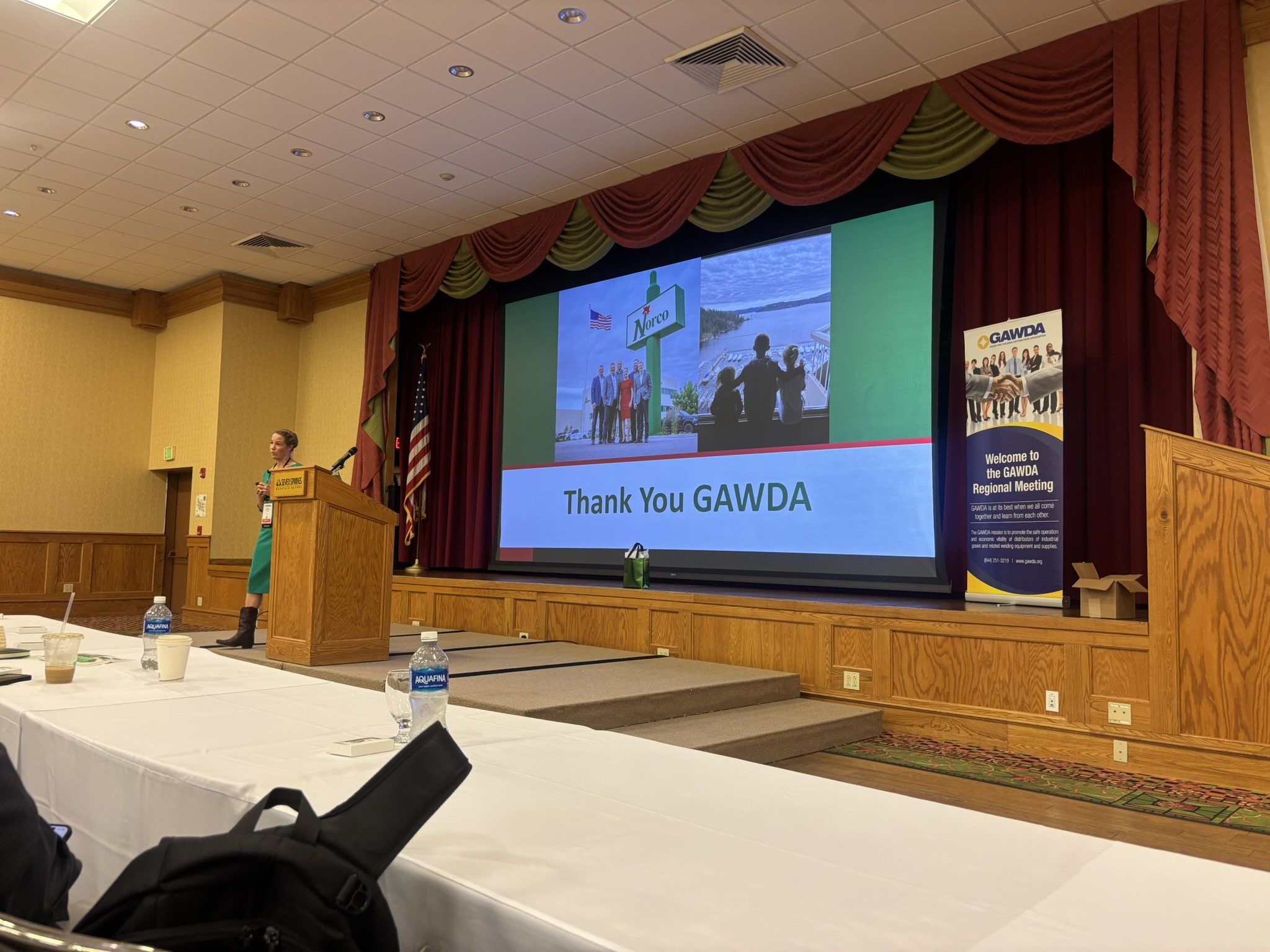 Speaker at a wooden podium giving a presentation in a conference room with a large screen reading 'Thank You GAWDA' and a banner nearby.