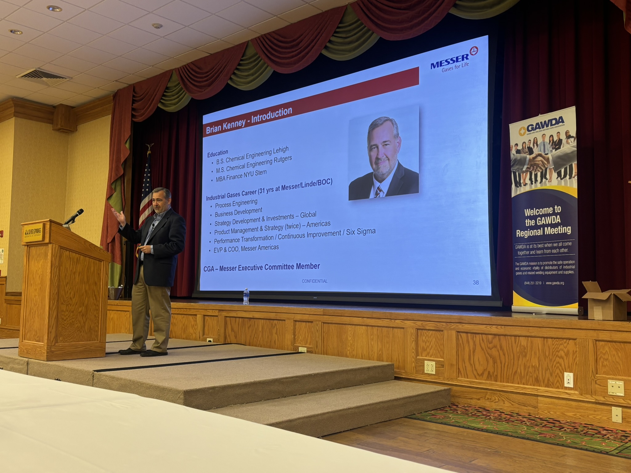 Speaker at a wooden podium presenting a slide titled 'Brian Kenney - Introduction' on a large projector screen behind him, in a conference room setting with red drapery.