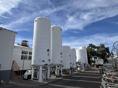 Industrial storage tanks lined up in an outdoor facility with a blue sky, white cylindrical tanks, and small safety placards visible on the sides.