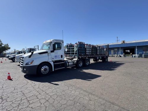 Line of white semi-trucks with flatbed trailers carrying stacked gas cylinders in a parking lot against a clear blue sky, with orange cones nearby.