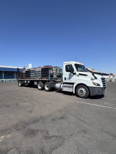White semi-truck with a flatbed carrying stacked gas cylinders in an industrial yard under a clear blue sky,