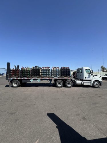 Flatbed truck carrying stacked gas cylinders on pallets in an open lot under a clear blue sky.