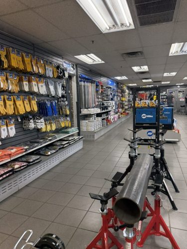 Hardware store interior with glove displays along the left, metal pipes and tools on shelves, and a large metal pipe on a red stand in the foreground.
