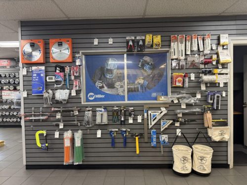 Wall display in a hardware store with welding and metalworking tools hanging on slatwall, including saw blades, clamps, files, measuring tapes, and miller welding curtain behind the display.