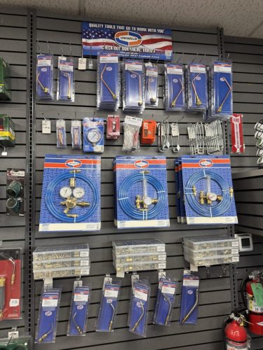 Wall display of welding accessories in blue packaging, including hoses and gauges, hanging on hooks in a hardware store.