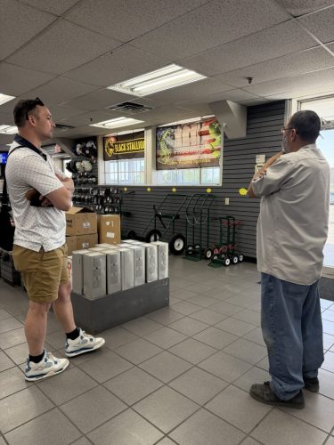 Two men stand facing each other in a store, arms crossed, mid-conversation among shelves of gear and banners.