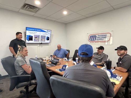 People sit around a conference table listening to a presentation; slide shows 'Transformer vs. Inverter' and a Phoenix Welding Supply banner on the wall.