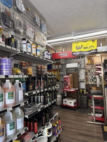 A hardware store aisle lined with shelves of cleaning and maintenance liquids, with a yellow Welder banner and a Lincoln Electric sign overhead.