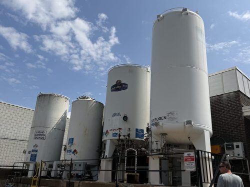 Row of tall white cryogenic gas tanks at an industrial facility, with 'Liquid Oxygen' and 'Nitrogen' labels, under a blue sky.