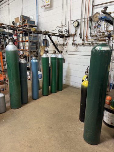 Row of tall green and blue gas cylinders standing upright in a workshop, connected to pipes and gauges along a white brick wall.