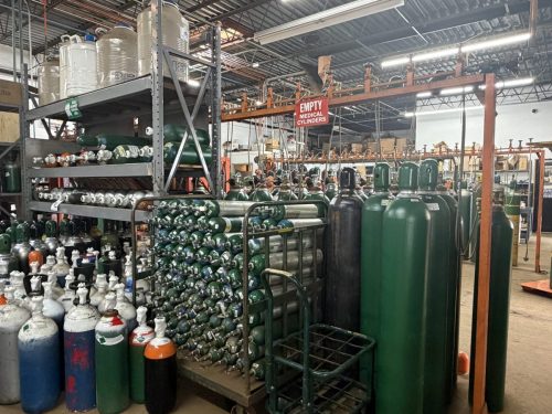 Industrial warehouse with rows of green and white gas cylinders on metal racks and a platform, sign reading 'EMPTY MEDICAL CYLINDERS' visible in the background.] ,