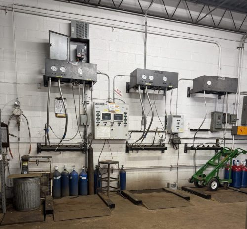 Industrial workshop wall with control panels, gauges, and exposed piping; blue gas cylinders lined along the floor and a green pallet jack on the right side.