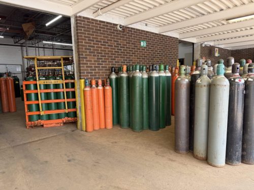 Row of large gas cylinders in green, orange, and gray lined up against a brick wall in an industrial storage area, with an orange pallet rack on the left.