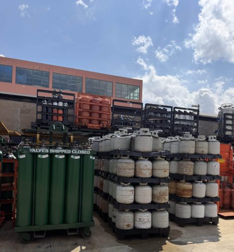 Outdoor storage yard with stacked propane/gas cylinders on pallets; pink building in background and blue sky above.
