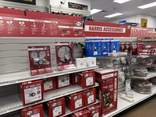 Store aisle with red Harris accessories display, shelves stocked with regulators, hoses, and gauges in red packaging.
