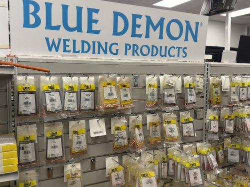 Store display with a large 'BLUE DEMON WELDING PRODUCTS' sign above rows of packaged welding accessories (TWECO) on hooks