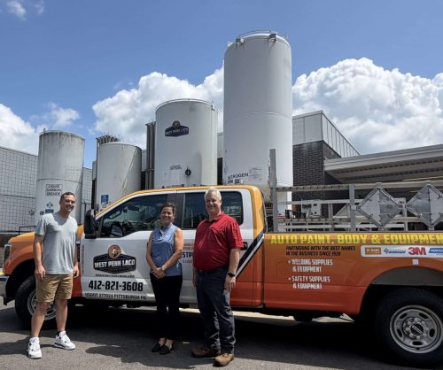 Three smiling people stand in front of an orange service van labeled 'Auto Paint, Body  Equipment' beside white industrial storage tanks under a blue sky.