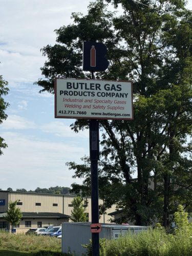 Outdoor Butler Gas sign on a tall pole with a blue B and a red gas cylinder icon, set against trees and a warehouse.