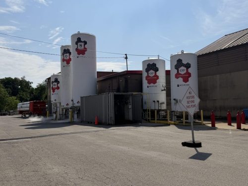 Industrial gas storage tanks in a yard beside a building under a blue sky, labeled CO2 and O2 with warning signs.