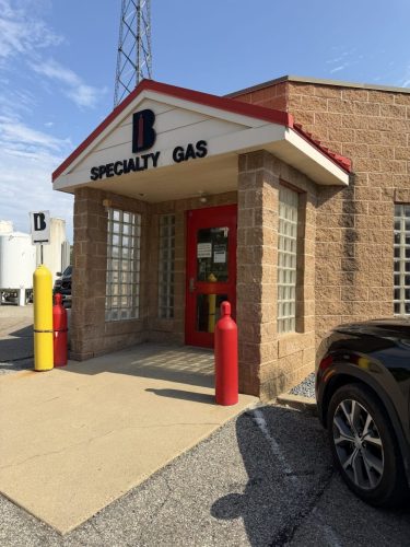 Gas station front with red door, beige stone walls, and a peaked white roof reading 'B Specialty Gas' under a clear blue sky; yellow and red safety bollards nearby.
