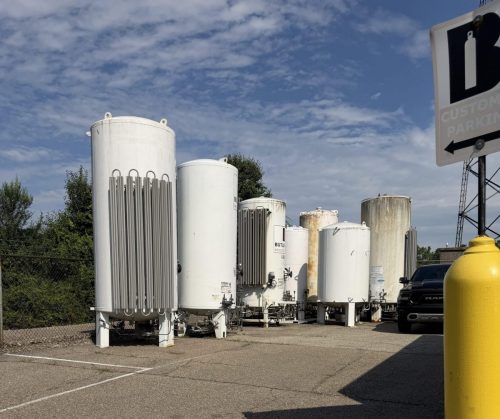 Row of tall white industrial storage tanks outdoors with a fence and a nearby yellow propane tank in the foreground, under a blue sky.