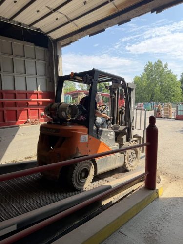 Orange Doosan forklift parked at a loading dock next to a red wall under an open garage door, ready for unloading.