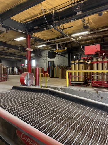 Industrial plant interior with rows of gas cylinders on racks, yellow railings, and exposed pipes under a damaged ceiling.