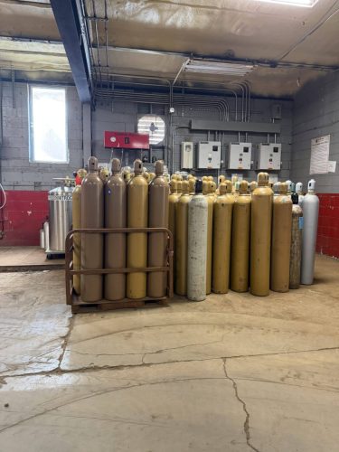 Row of tall gas cylinders in a metal rack inside an industrial utility room with exposed pipes and a concrete floor.