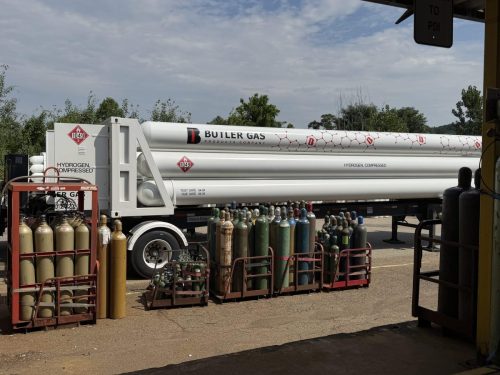 Industrial gas yard with a white Butler Gas trailer and racks of compressed gas cylinders in foreground, outdoors under a cloudy sky.