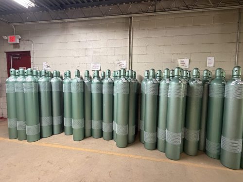 Row of tall green gas cylinders with protective wrap, lined up against a concrete wall in a warehouse area.