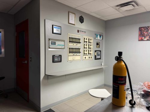 Industrial control panel wall with gauges and switches, certificates above, and a yellow gas cylinder in the foreground.
