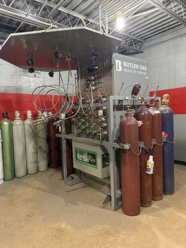 Industrial gas storage and distribution system with tall gas cylinders connected by hoses and fittings, labeled Butler Gas Products Company on a gray equipment rack.