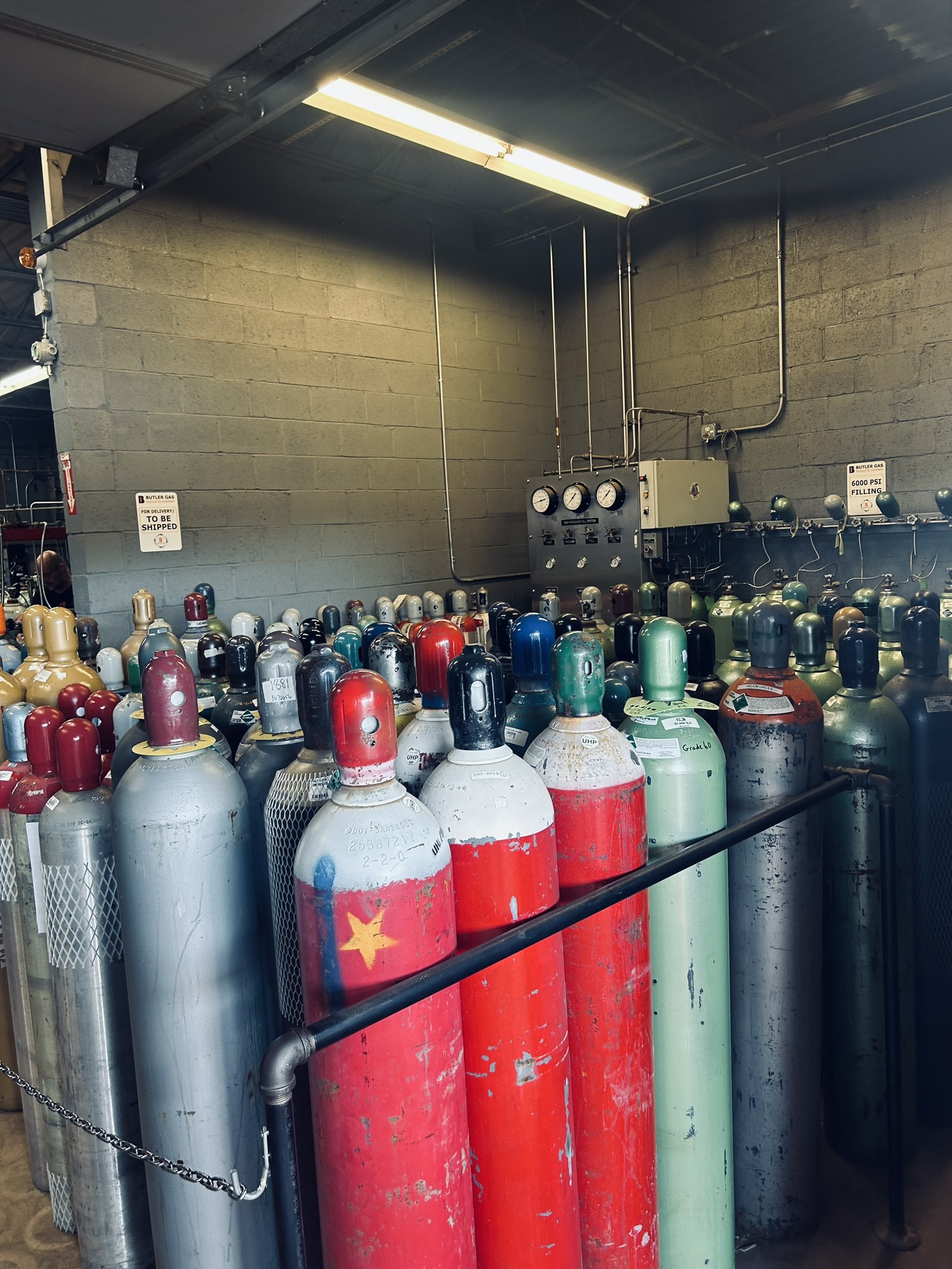 Row of colorful gas cylinders in an industrial storage area, with pipes and a control panel on the back wall.
