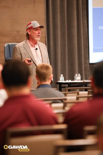 Speaker in a gray blazer and pink shirt presenting to an audience, wearing a red-and-gray cap and holding a clicker with a microphone clipped to his lapel.