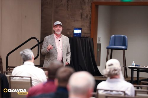 Male speaker in blazer and cap presenting to an audience at a conference, standing on a stage with a laptop on a tall table behind him.